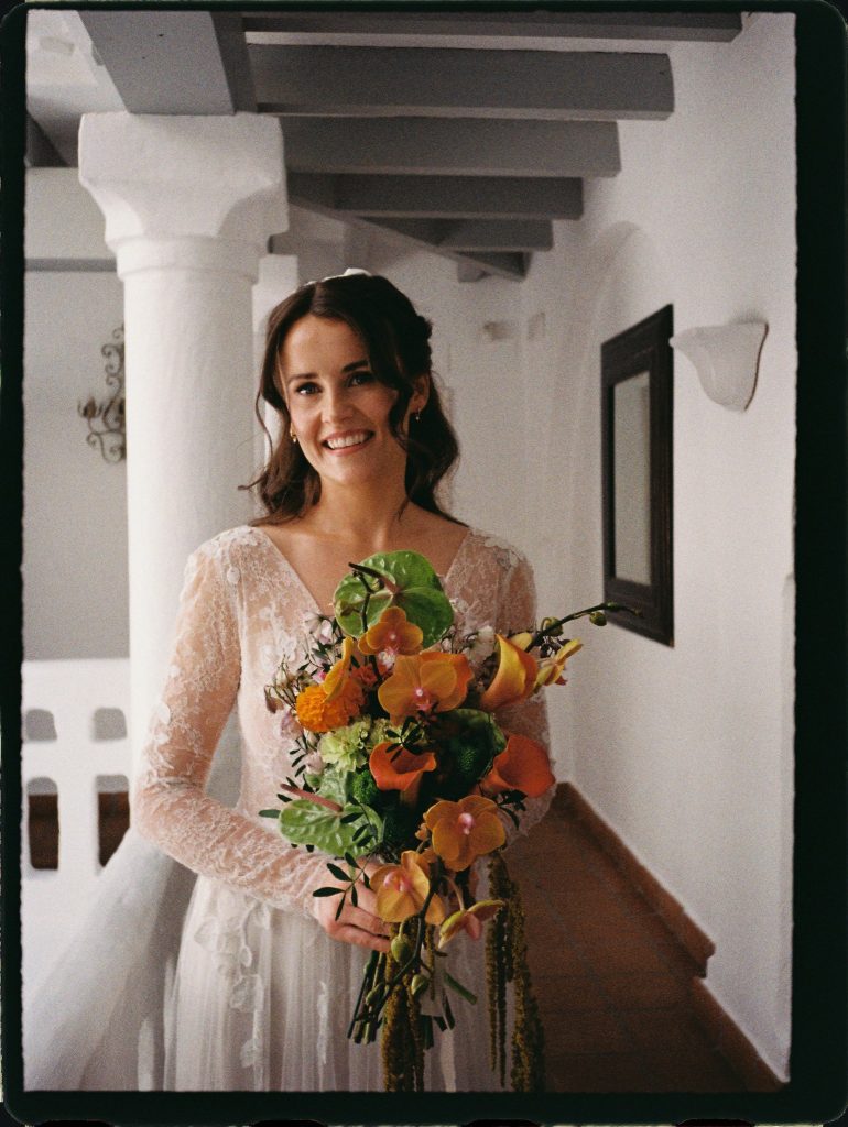 Smiling bride with an orange orchid bouquet in a white hallway, 35mm film wedding photo on the Costa del Sol