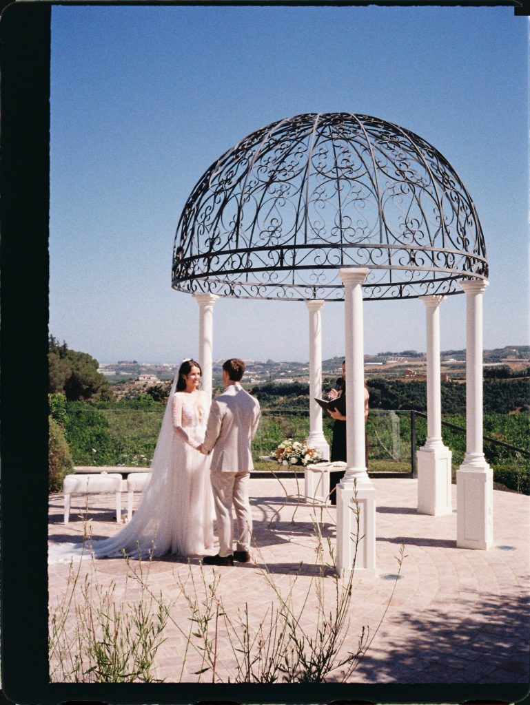Ceremony under a wrought-iron gazebo with views of the coast, 35mm film wedding photo on the Costa del Sol