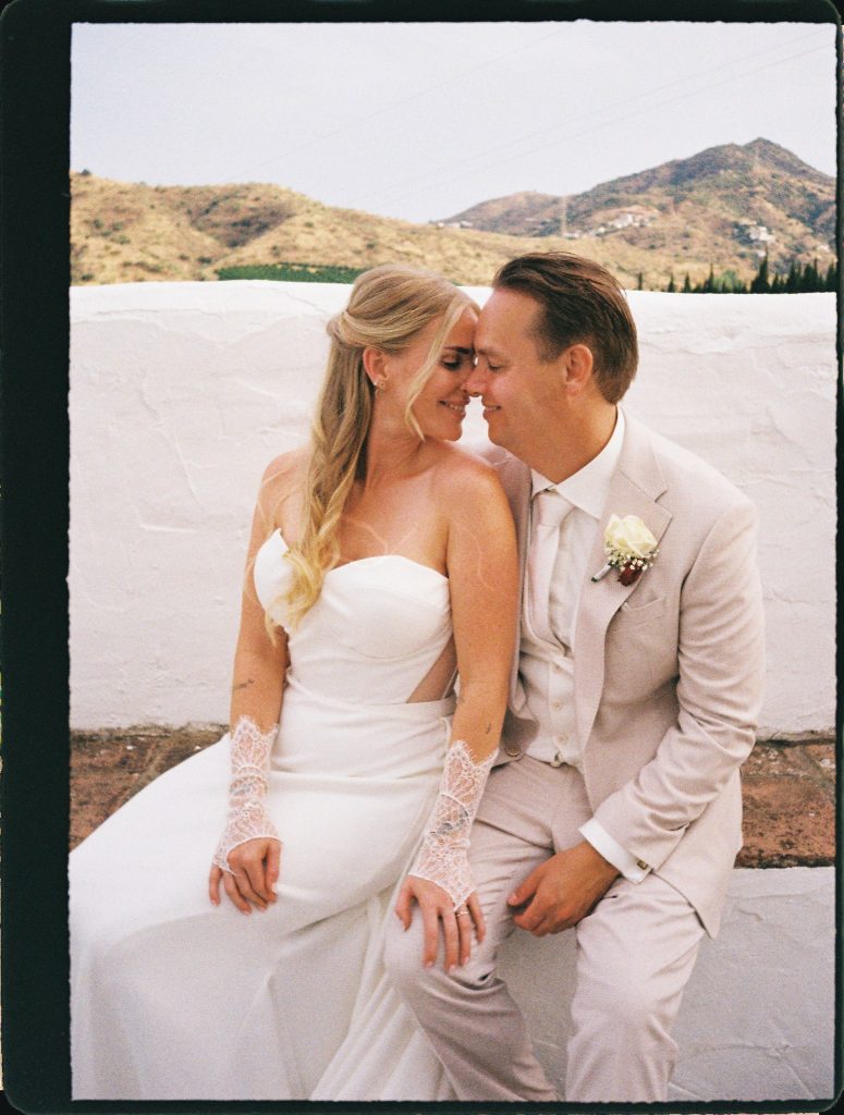 Bride and groom touching foreheads by a white wall with mountains behind, 35mm film wedding photo on the Costa del Sol